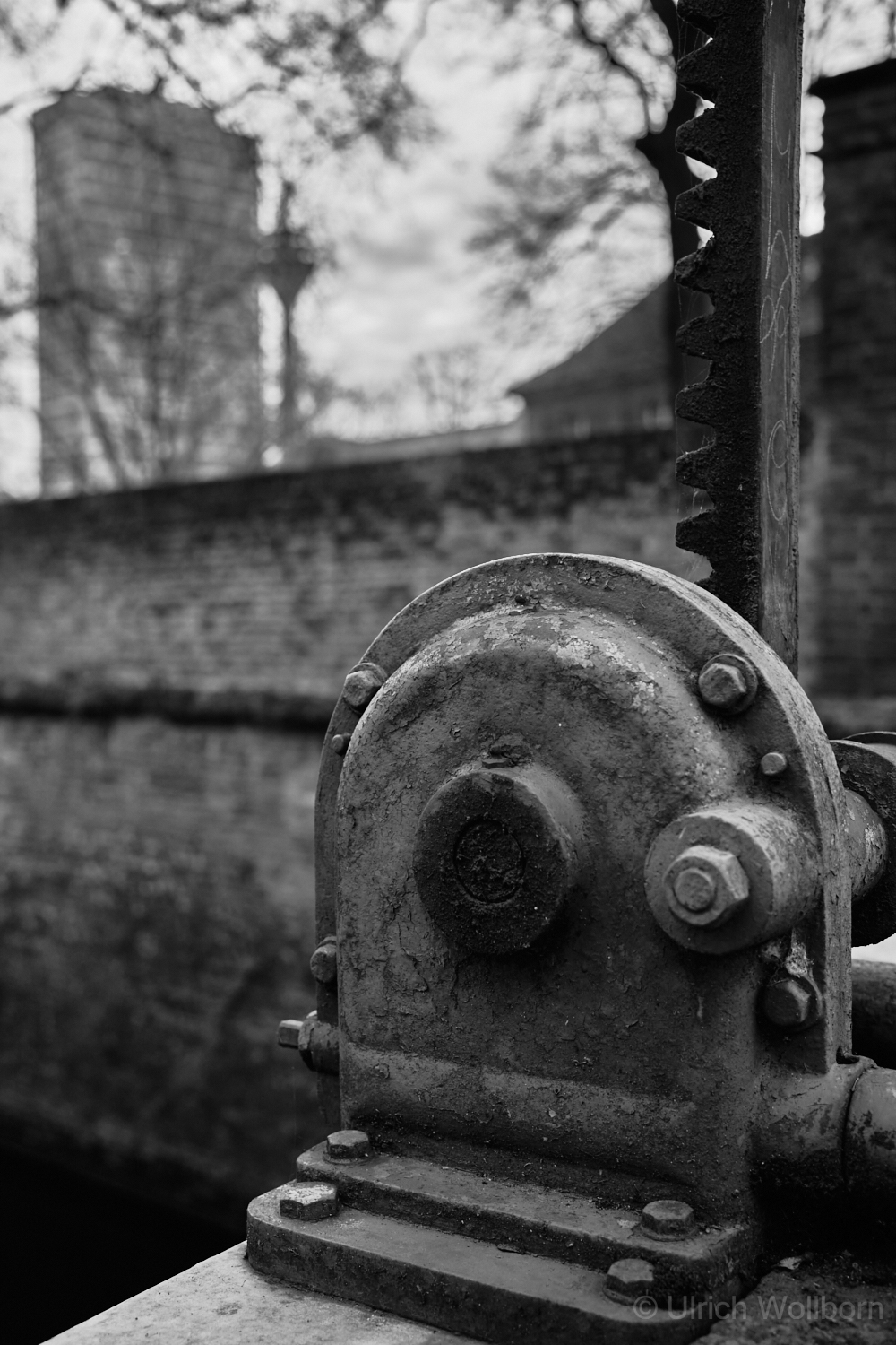 Black-and-white photograph showing rusty mechanical parts of the Düsseldorf Spee'scher Graben water control structure in the foreground with the Mannesmann Tower and Düsseldorf TV Tower blurred in the background.