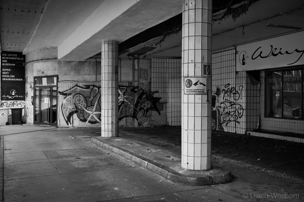 Black and white photo of an old, abandoned gas station featuring tiled columns and walls covered with graffiti, peeling paint, and a worn entrance door. The site shows signs of neglect and urban decay, with scattered debris and faded signage.
