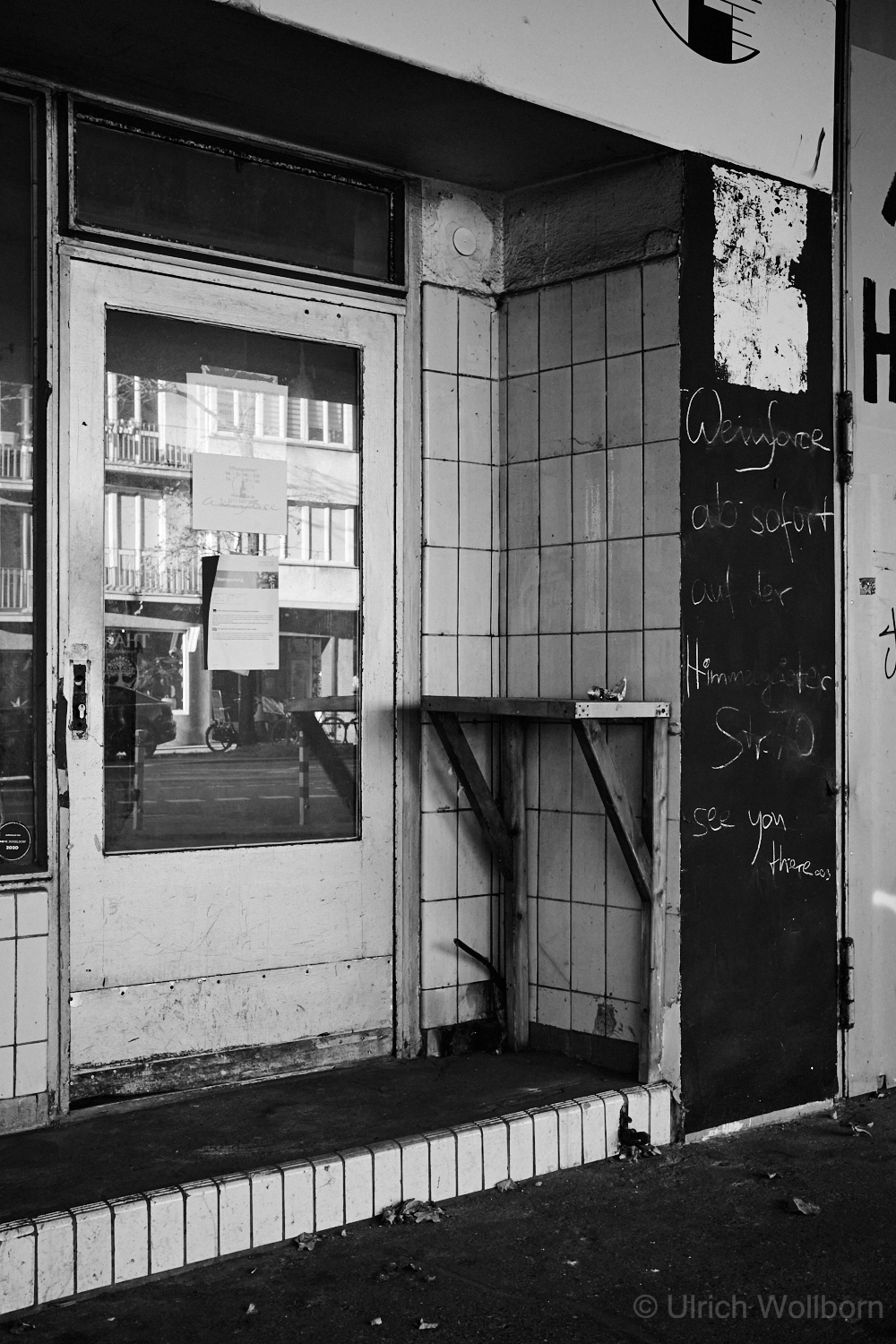 Black and white photo of an old, abandoned gas station featuring tiled columns and walls covered with graffiti, peeling paint, and a worn entrance door. The site shows signs of neglect and urban decay, with scattered debris and faded signage.