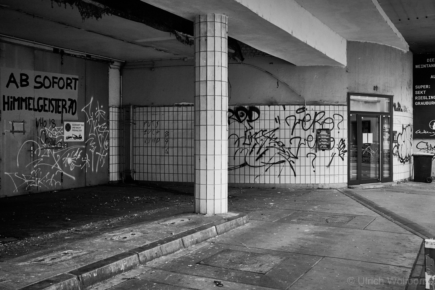 Black and white photo of an old, abandoned gas station featuring tiled columns and walls covered with graffiti, peeling paint, and a worn entrance door. The site shows signs of neglect and urban decay, with scattered debris and faded signage.