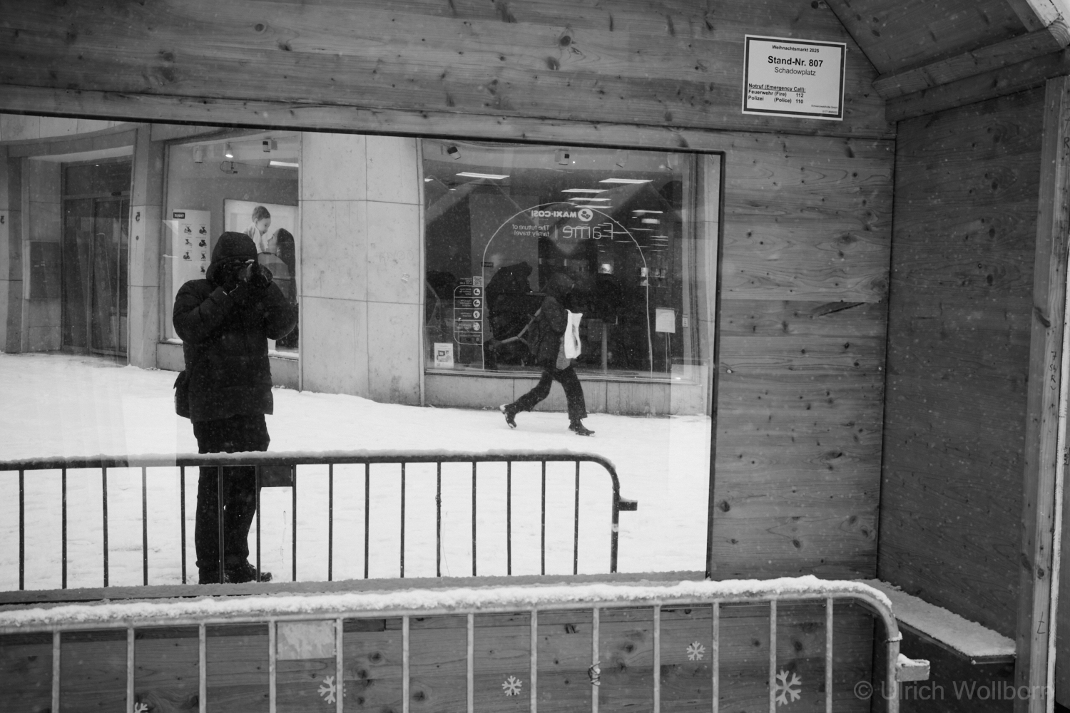Black and white photo showing me, dressed in winter clothing, taking a photograph through the reflective window of a wooden Christmas market booth that is being dismantled. Snow covers the ground outside, and another person walks past a storefront in the background. The scene captures a cold, urban environment with metal barriers in the foreground.