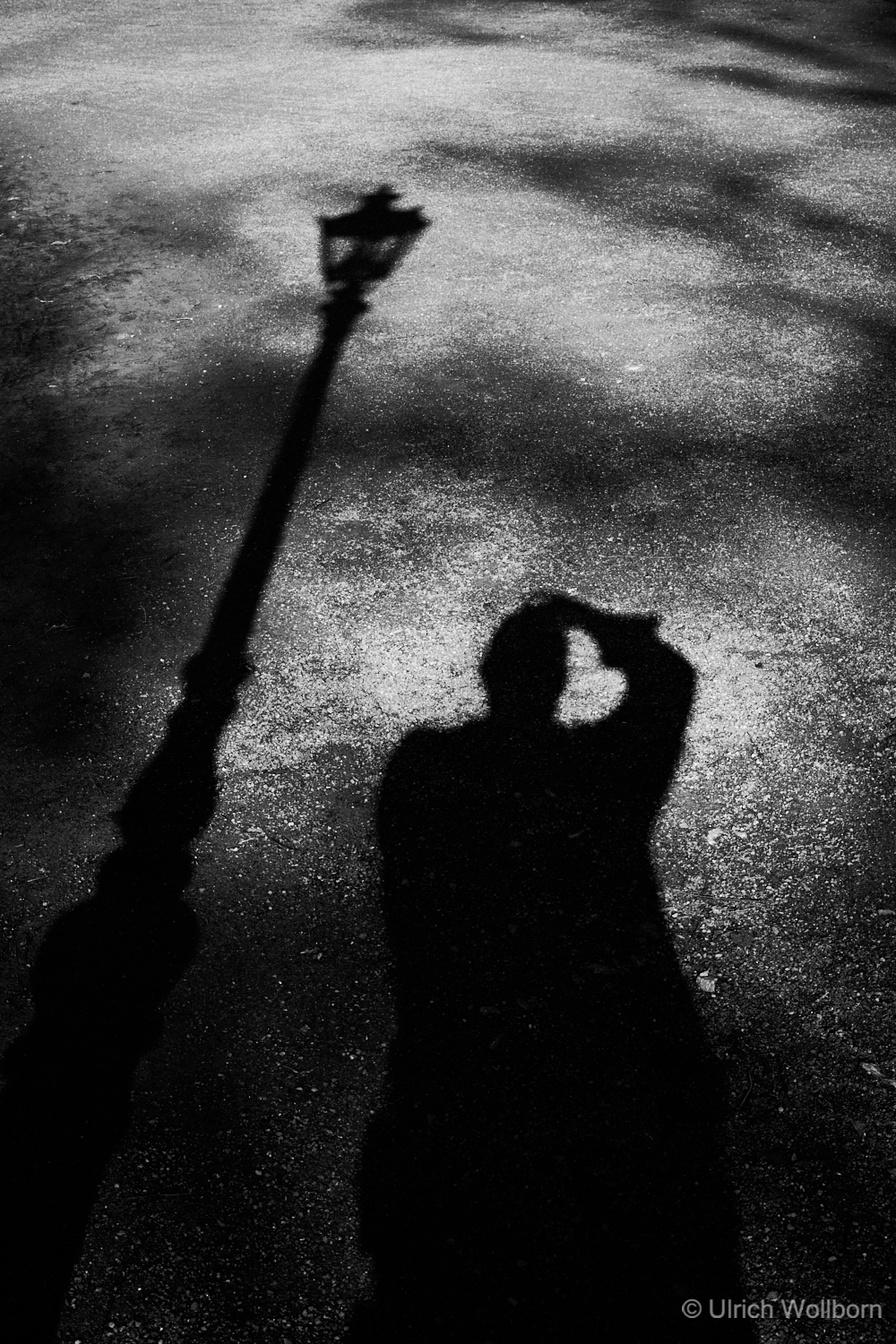 Black-and-white shadow photograph: The long shadow of a historic street lamp and the shadow of the photographer Ulrich Wollborn are visible on the ground.