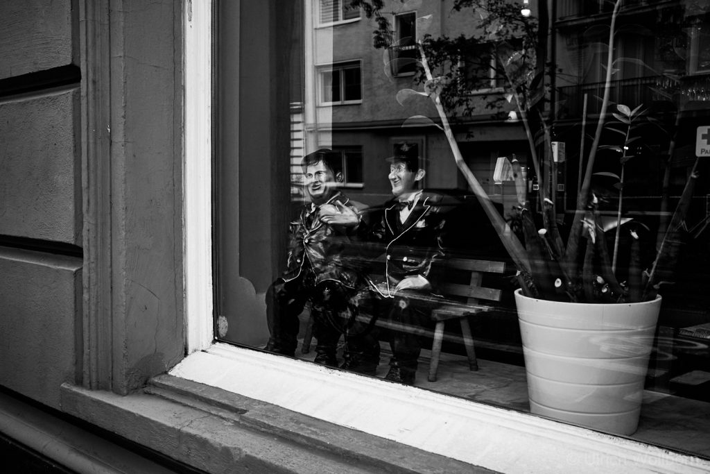 Black and white photo looking through a shop window, showing two mannequins of the famous comedy duo 'Laurel and Hardy' (Stan Laurel and Oliver Hardy) dressed in formal suits, sitting on a bench. One mannequin is gesturing. Reflections of buildings, trees, and the street are visible in the window, along with a potted plant on the right.