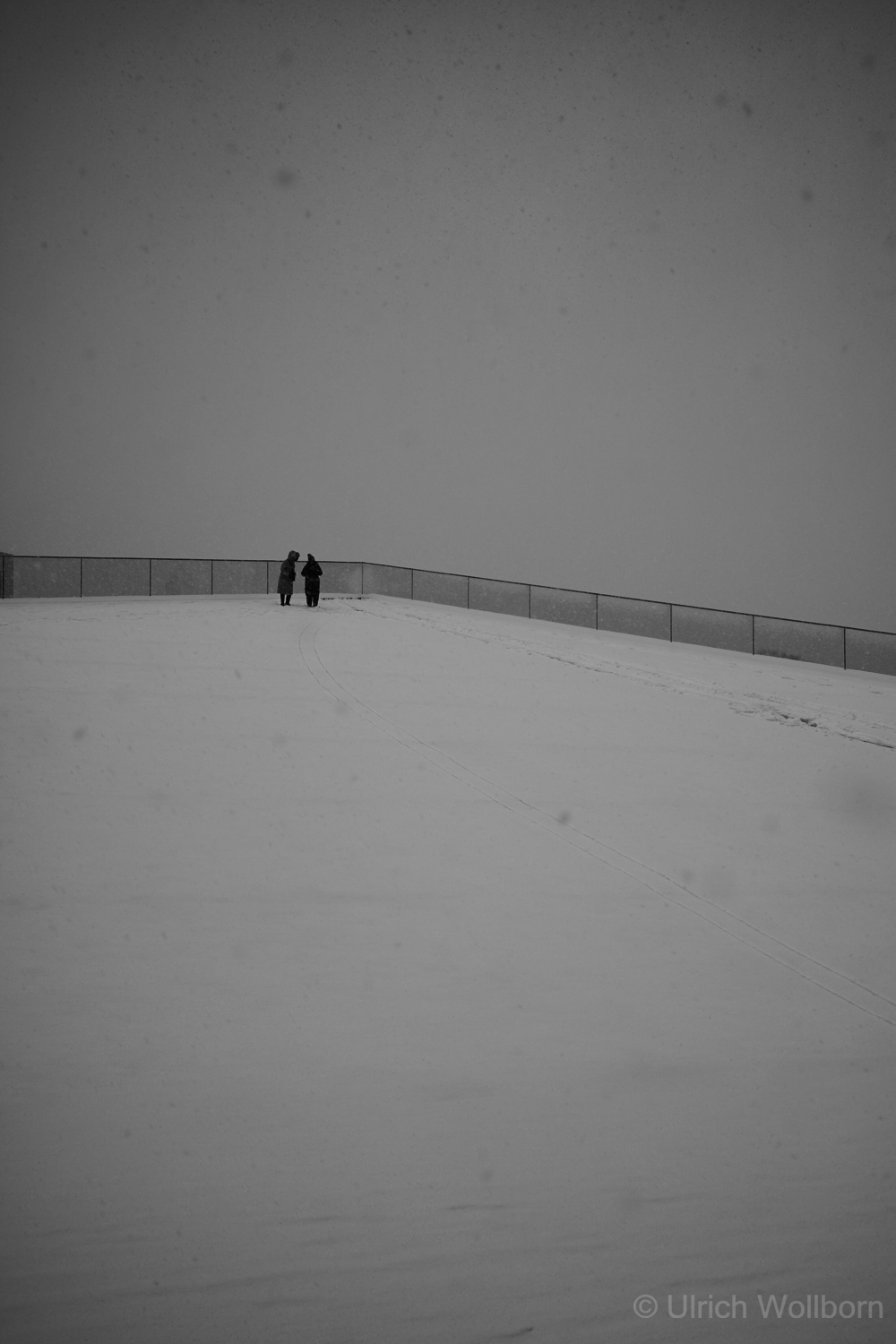 Two people stand on a snow-covered hill next to a fence under a gray, overcast sky.