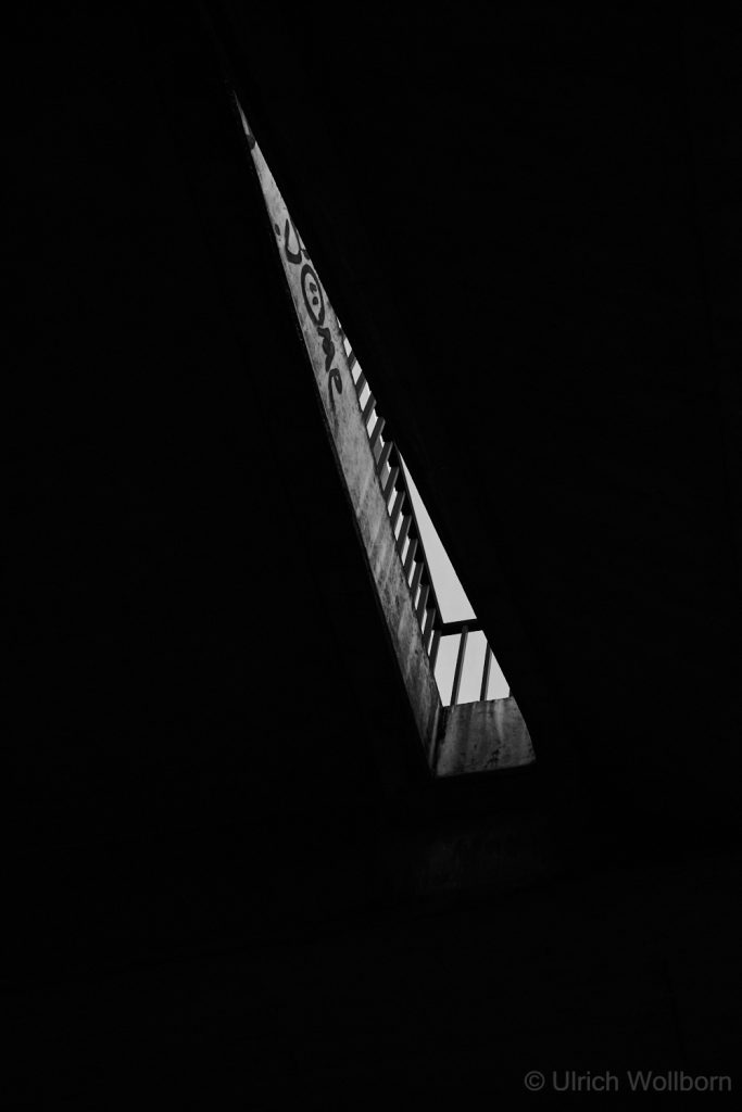 A black and white photo showing a narrow vertical opening between dark urban structures, revealing a sliver of sky and a railing. The composition highlights strong contrasts, geometric lines, and the interplay of light and shadow in an urban setting.