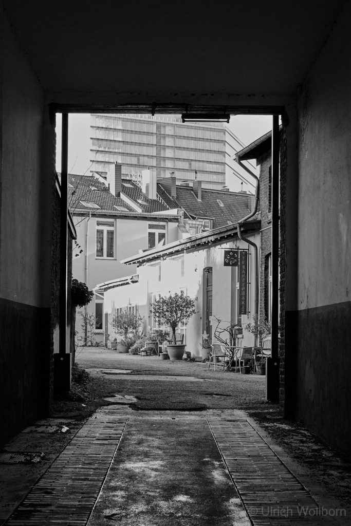 Black and white photo looking through a dark alleyway opening onto a bright courtyard with potted plants, outdoor chairs, and a mix of old and modern buildings in the background.