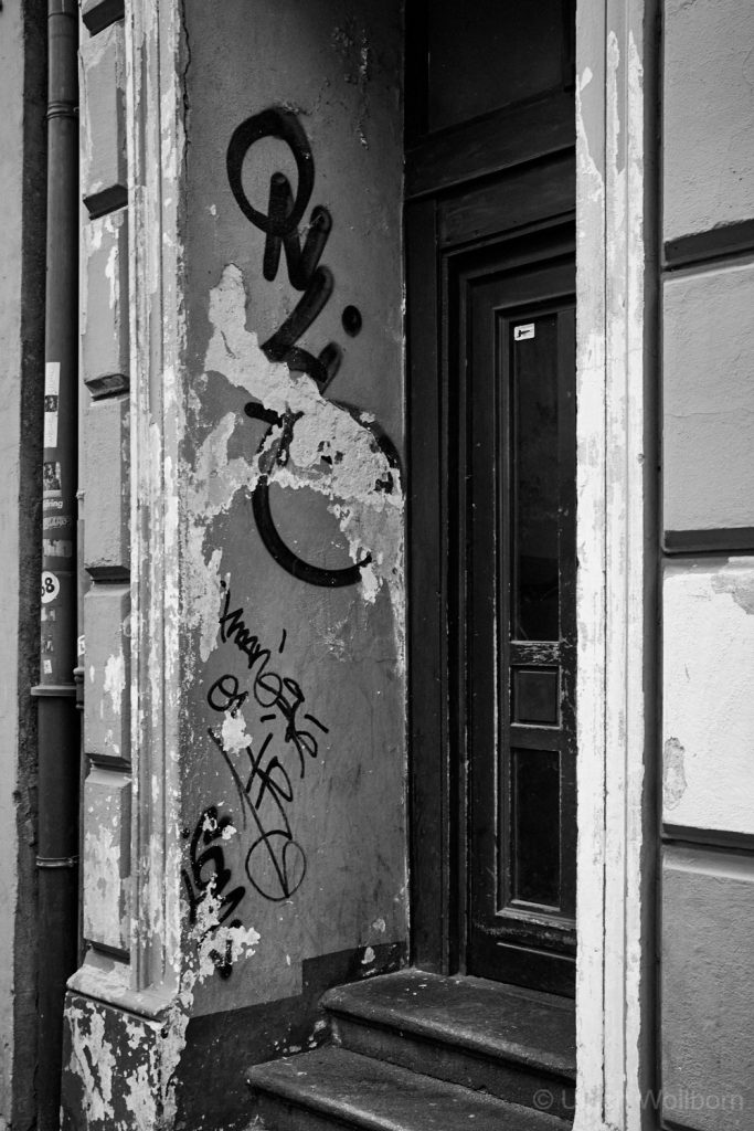 Black and white photo of an old urban doorway with peeling paint on the walls, graffiti tags on the left side, and a dark wooden door with steps leading up to it.