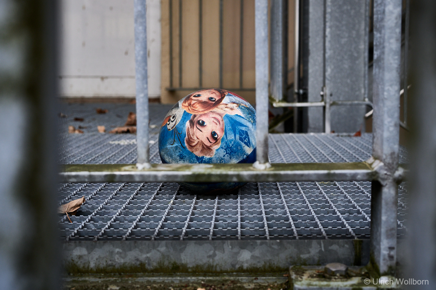 Vividly colored cartoon-themed ball resting on a metal fire escape staircase, framed by cold steel bars and scattered autumn leaves, evoking contrast between innocence and industrial surroundings.