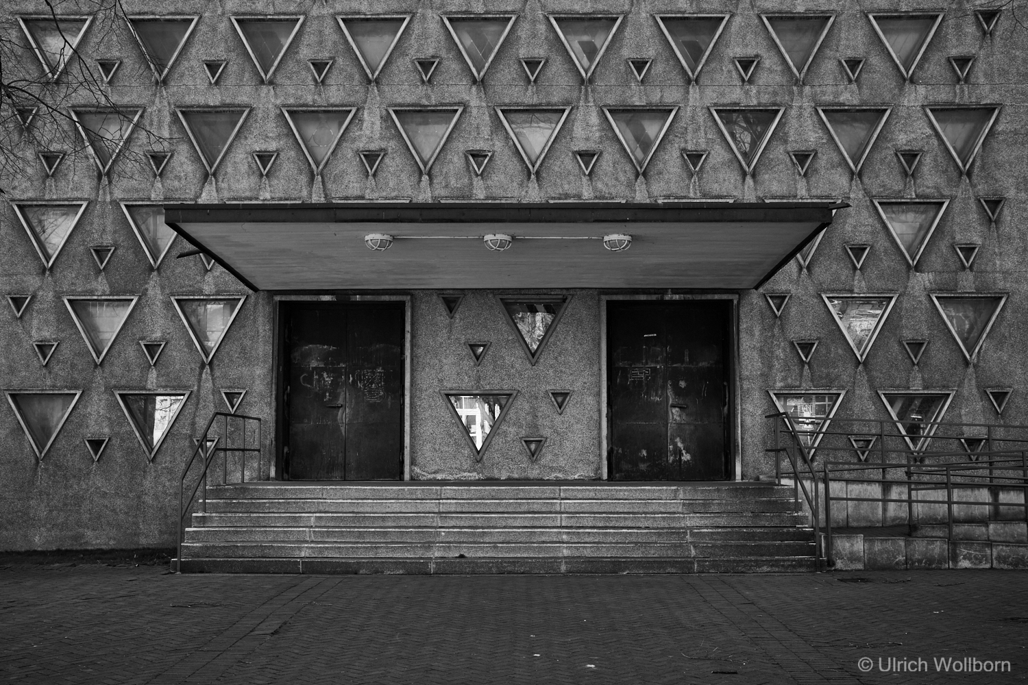 Black and white image showing the front entrance of an abandoned building with a unique facade composed of numerous triangular windows. Two dark doors are centered, framed by concrete stairs and metal railings.
