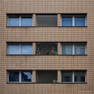 Minimalist facade of a beige apartment block with geometric window patterns, featuring a single balcony adorned with vibrant flowers and hanging decorations, highlighting human presence in urban order.