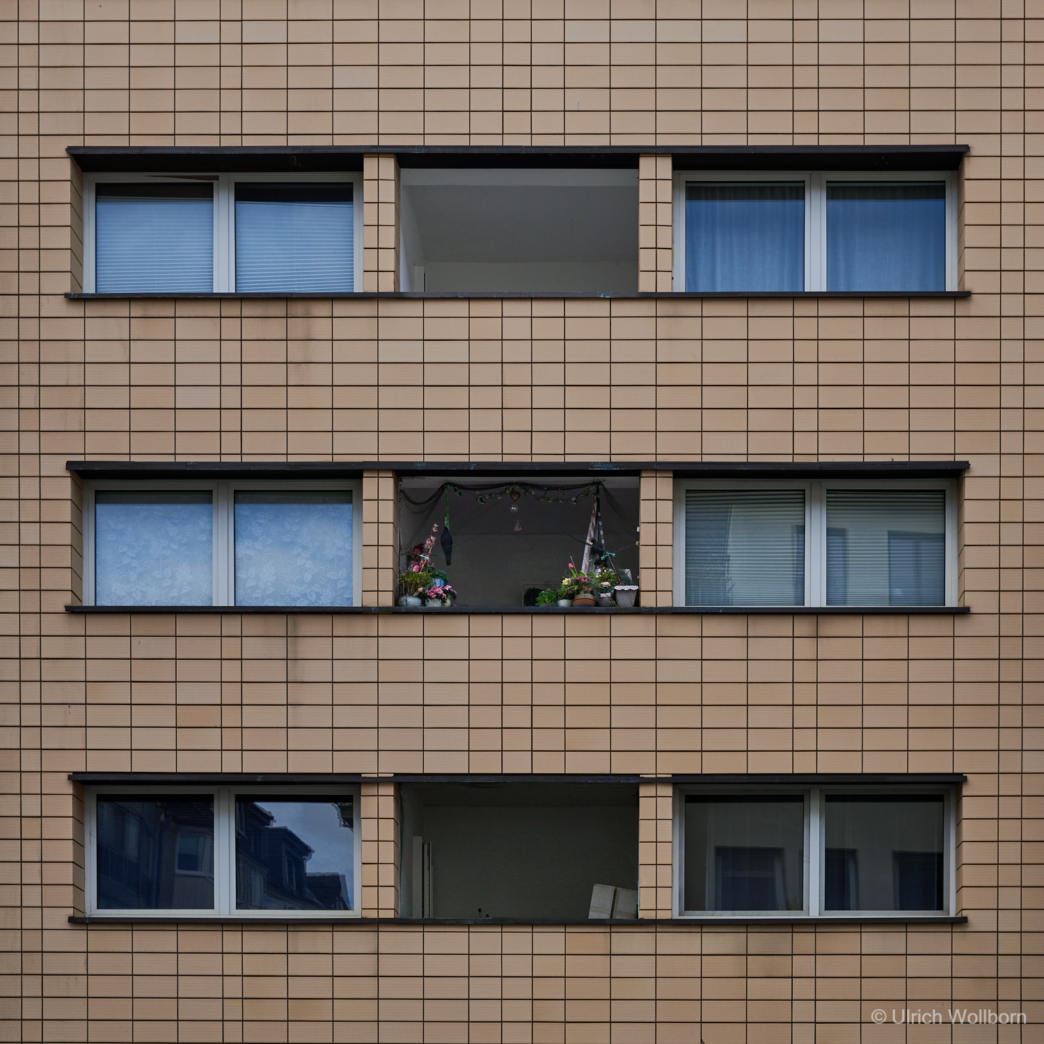 Minimalist facade of a beige apartment block with geometric window patterns, featuring a single balcony adorned with vibrant flowers and hanging decorations, highlighting human presence in urban order.