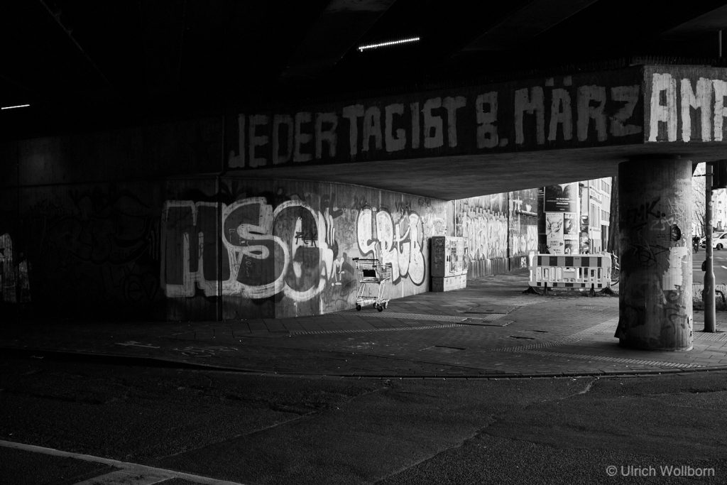 Black and white photo of an urban underpass covered in graffiti, featuring a lone shopping cart against the wall and a concrete pillar. The graffiti includes large letters and a German phrase painted on the overhead beam.