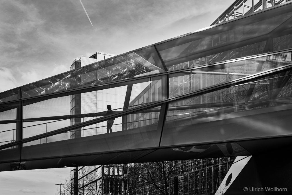 Silhouette of a person walking inside a modern glass pedestrian bridge with urban buildings in the background, captured in black and white.