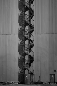 Black and white photograph of a spiraling metal staircase ascending vertically along a corrugated gray industrial wall, with a door to the hall at the base and a no-entry sign on the right.