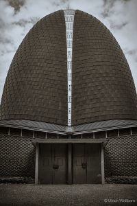 Close-up view of a large, dome-shaped building with a textured, shingle-like exterior and a vertical strip of metal panels running down the center. The structure features two large, dark metal doors at the base, set against a patterned wall under a cloudy sky.