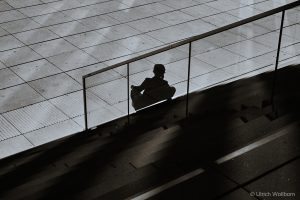 Black and white photo showing the shadow of a person sitting on stairs, reflected on a glass railing. The background features a tiled pavement with subtle reflections, creating a contemplative and quiet urban atmosphere.