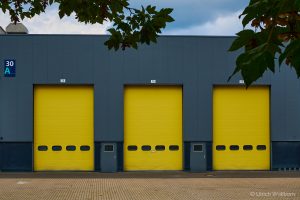 Three vibrant yellow industrial rolling doors numbered 73, 74, and 75 on a gray warehouse facade marked section 30 A, framed by overhanging tree branches against a partly cloudy sky.